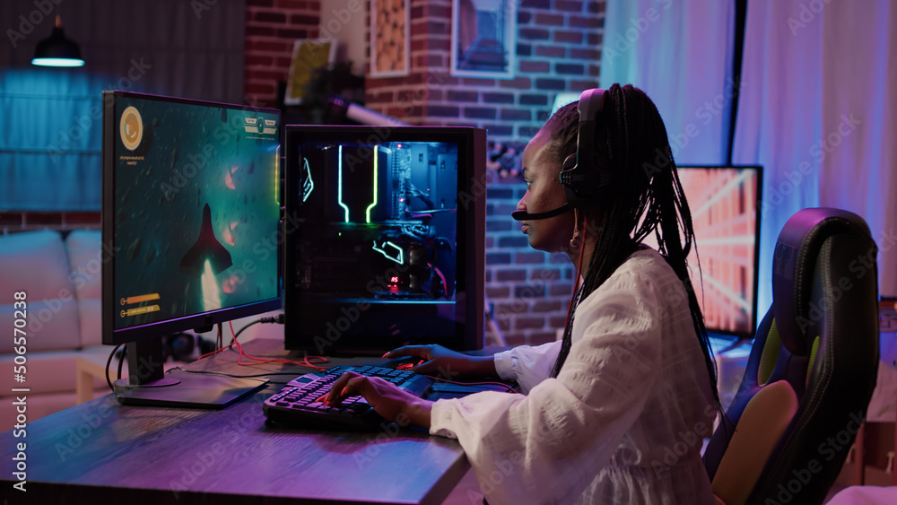 Wall mural African american gamer girl using pc gaming setup playing multiplayer space shooter simulation having a good time in home living room. Woman streaming online action game while talking in headset.