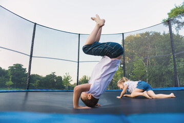 A boy and a girl jump on a trampoline without parental supervision. Brother and sister play on the trampoline in the park in summer - obrazy, fototapety, plakaty