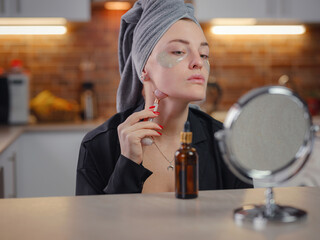 Sculpting, lifting and anti-aging care. Skin care and beauty at home. Woman using calming quartz roller massager while sitting on sofa in kitchen looking in mirror. Woman using face roller.