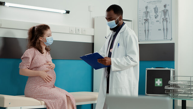 Pregnant Woman Talking To Doctor About Pregnancy At Checkup Visit During Covid 19 Pandemic. Male Physician Doing Health Consultation With Patient Expecting Child In Medical Cabinet.