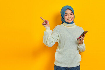 Fototapeta na wymiar Portrait of smiling young asian woman in white sweater and hijab holding mobile phone, pointing finger aside isolated over yellow background. People islam religious concept