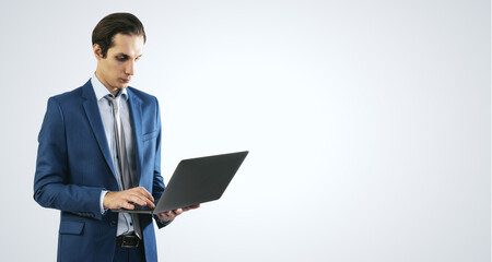Young businessman in blue suit working with modern laptop, isolated on light grey wall background,...