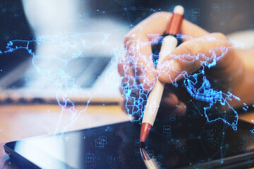 Close up of hands using digital tablet with pen and laptop with glowing blue map hologram and digital data concept on blurry office desktop background. Info, news and planet concept. Double exposure.