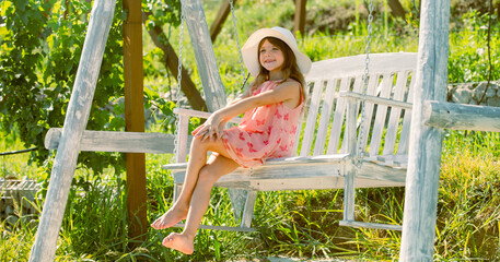 Happy little child girl on a swing in the summer park. Beautiful child girl in hat dreaming. Fashion child in summer straw hat and sunglasses. Dreamy kids face. Child chill and resting in summer