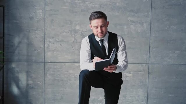 Young business man Caucasian nationality with beard, middle aged, classic clothes, white shirt black trousers, poses interior office with notebook his hands, speaks writes with pen. Businessman