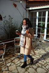 A freelancer walks in front of the cafe with coffee to go and a laptop in her hands.