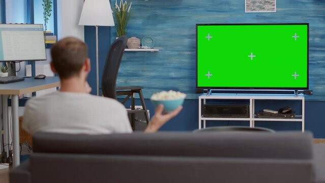 Man relaxing on couch looking at green screen on tv watching movie while eating popcorn in modern living room. Person having a good time sitting on sofa looking at television with chroma key display.