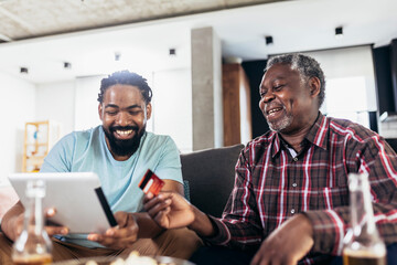 Smiling father and son using digital tablet and credit card for online payment in living room at...