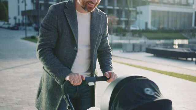 At sunlight handsome father with little new born baby in a stroller plays smiling. In city at sunlight on street. Outdoors. Slow motion
