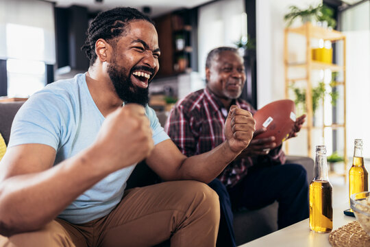 Senior Father And Adult Son Cheering For American Football Game At Home