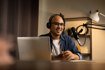 Young asian man host streaming podcast with condenser microphone work on laptop at small broadcast home studio. Content creator blogger recording voice over radio interview guest conversation