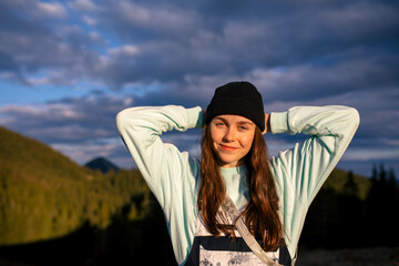 Incredible girl in a black hat stands in the mountains with arms raised against the backdrop of mountains and with a very beautiful sunset