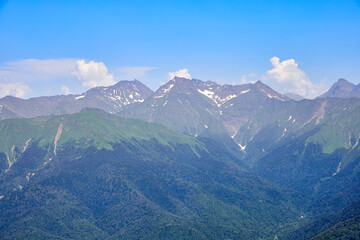 Fototapeta premium Mountain bright landscape with snow-capped mountains, blue sky and clouds. Mountainside covered with green grass and fir trees.