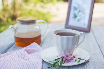 Glass teapot with tea and a white ceramic cup on a wooden table. In the background are flowers in a white jug and a painting.