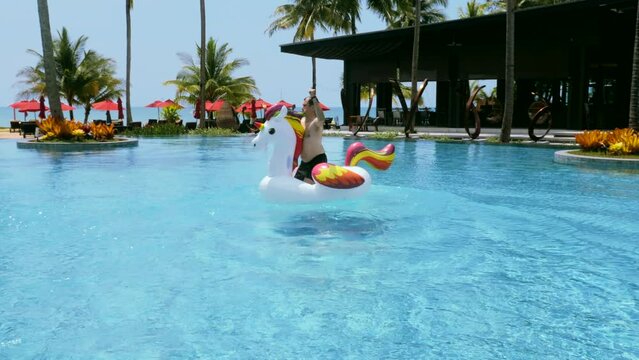 Happy adult man having fun, ride and dancing on floating inflatable unicorn in swimming pool in luxury hotel. Funny male tourist enjoying summer holidays, vacations, sunny day, sea view on background.