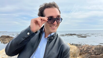 A handsome French man in black sunglasses stands on the shores of the Atlantic Ocean, he has black hair, a leather jacket and he flies with his hands to the sides travel advertising High quality photo