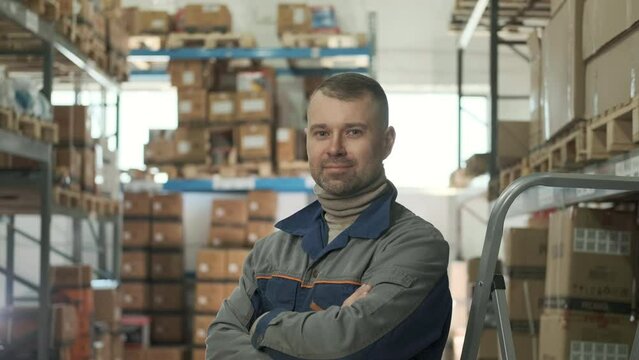 Professional worker in overalls smiles happily at camera. In background is large warehouse with shelves full of goods for delivery. Portrait of container storage warehouse employee