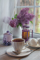 Obraz na płótnie Canvas Fresh, fragrant tea in a white ceramic cup, marshmallows on a white plate. In the background against the background of the window is a bouquet of lilacs in a white jug.