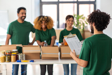 charity, donation and volunteering concept - international group of happy smiling volunteers packing food in boxes according to list on clipboard at distribution or refugee assistance center