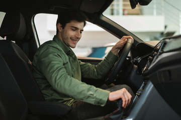 Happy young Caucasian man test driving new car before purchase at modern automobile dealership,...