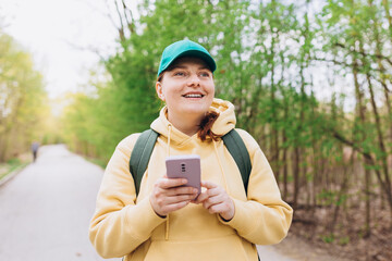 A portrait of a smiling beautiful woman in green cap texting sms with her phone on nature background. Happy woman with backpack is using a smartphone in park outdoors, spring time. Traveler