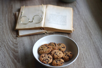 Bowl of strawberries and blueberries, open books with reading glasses, plate of chocolate chip cookies and basket of apples one the table. Selective focus.