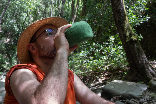 Hombre bebiendo en cantimplora en medio del bosque con sombrero de paja, excursi&oacute;n de vacaciones, vegetaci&oacute;n