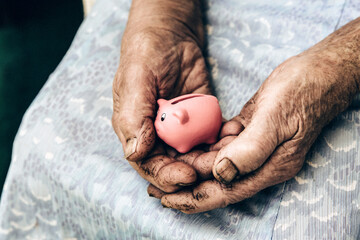 Close up view of old rough elderly retired woman hands holding tiny pink piggy bank. Poverty and...
