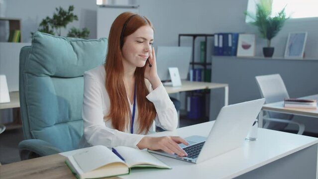 Smiling girl in call center. Happy young woman in headset using laptop in modern office, office worker laughing, talking on cell phone with client, using wireless internet connection. Indoors, daytime