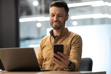 Happy Businessman Holding Cellphone Texting Working On Laptop In Office