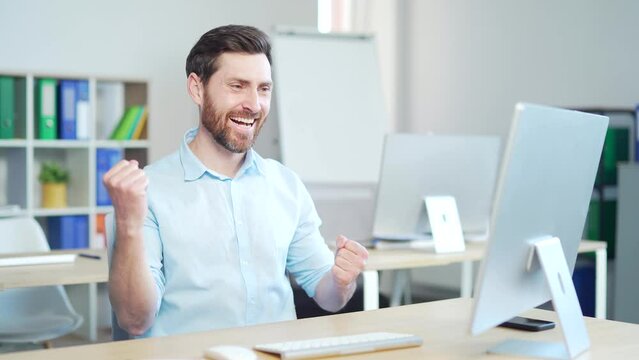 Happy Handsome bearded Business Man Employee Working on pc computer Celebrates Successful Endeavor with YES Gesture. Working in moderm office Has Stroke of Luck, Wins Big. worker victory good news