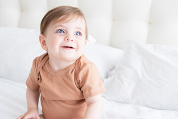 portrait of cute baby girl 10 months with blue eyes in brown bodysuit on white bedding on bed - obrazy, fototapety, plakaty