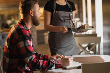 female waitress giving order to man, talk to client cafe restaurant visitor, friendly professional woman server wearing apron write lunch or breakfast food menu selection, serving staff