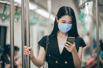 Young Asian businesswoman with protective face using smartphone while commuting in the city riding...