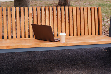 A gray laptop and a cup of coffee on a wooden bench in the park. Freelancer's place of work. Study and work online
