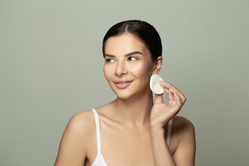 Smiling attractive woman cleaning her face with a cotton pad on gray background