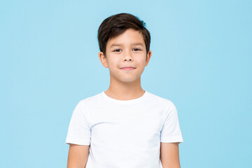 Cute smiling boy in plain white t shirt looking at camera in isolated studio light blue color background - obrazy, fototapety, plakaty