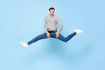 Fun energetic young Caucasian man wearing headphones listening to music and jumping with spread legs in studio light blue color isolated background