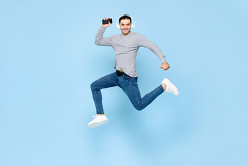 Cheerful young Caucasian man wearing headphones listening to music from smartphone and jumping in studio light blue color isolated background