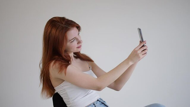 Beautiful young woman takes a selfie photo on the phone. Pretty female use selfie cam. Woman scratches her nose, looks at the resulting photos on the phone.