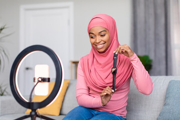 Excited muslim lady blogger recording video review of new smartwatch model, using ring lamp and smartphone at home - obrazy, fototapety, plakaty