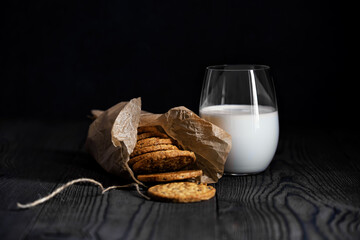 A glass of milk and muesli biscuits in a paper bag on a dark background. The wood texture of the...