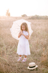 A cute little girl with long blond curly hair in a white summer dress with lace umbrella  in a field in the countryside in summer at sunset. Nature and Ecolife