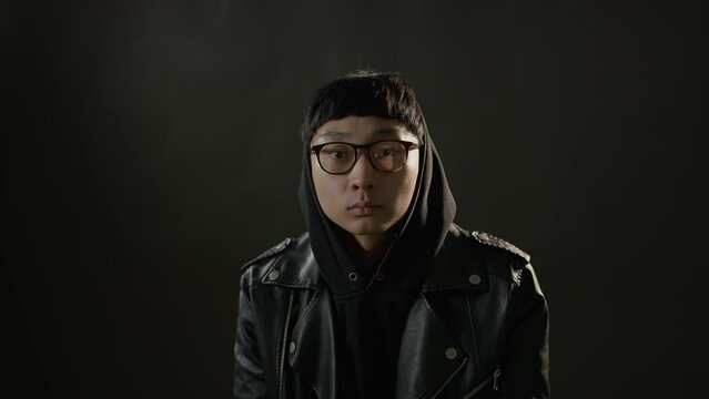A man puts in glasses and a hood turn around itself. Shot from the film. Man looks at the camera against a dark background. 