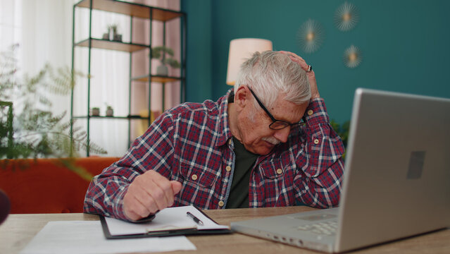 Unhappy Senior Old Grandfather Man At Home Living Room Working On Laptop Computer, Making Notes On Sheet Of Paper, Upset By Poor Results, Grabs His Head. Modern Technology For Retirement Pensioners