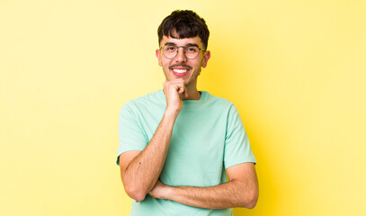 young adult hispanic man looking happy and smiling with hand on chin, wondering or asking a question, comparing options