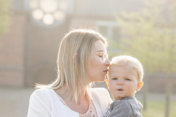 a young happy mother with a small child in her arms, gently kissing his head, stands in the middle of a park in the rays of the rising morning sun. High quality photo