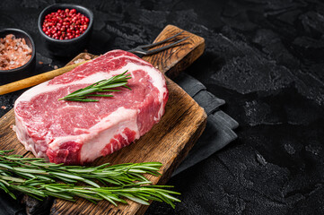 Raw Rib-eye Steak, beef marbled meat on wooden board with rosemary. Black background. Top view. Copy space