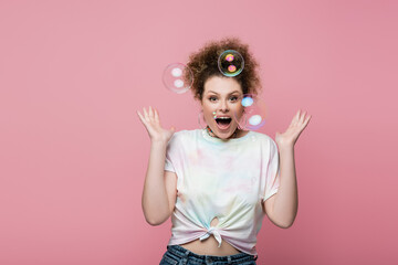 Shocked young woman standing near soap bubbles on pink background.