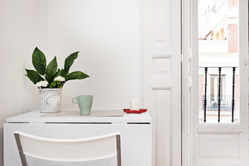 Studio apartment with white folding dining table with faux vase, candle in red ashtray, balcony with vintage white wooden windows and shutters and matching folding chairs